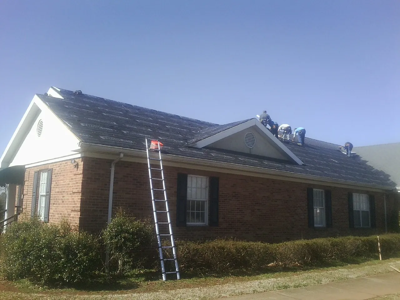 Crew tearing shingles off a brick ranch roof with a ladder set — Emergency Roof Repair work by our crew in Benedict