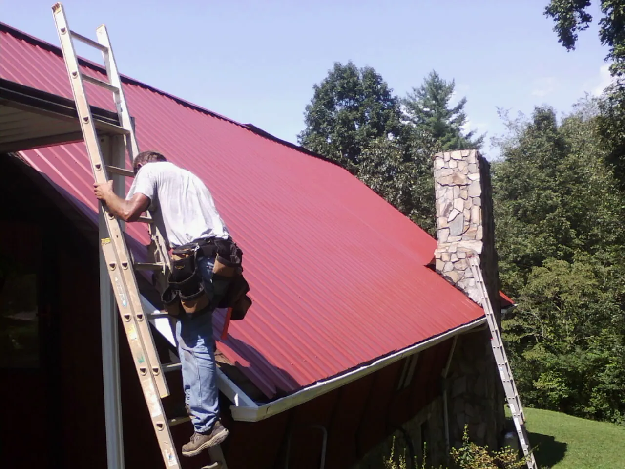 Roofer on a ladder beside a red metal roof and stone chimney — Soffit & Fascia Repair by Roofing Brothers in Benedict